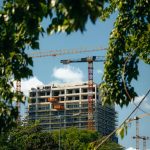 High-rise building under construction with cranes in Bratislava, framed by greenery.